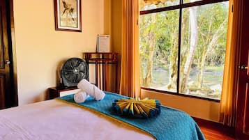 A hotel room with a bed, a fan, a window with curtains, and a framed picture on the wall.