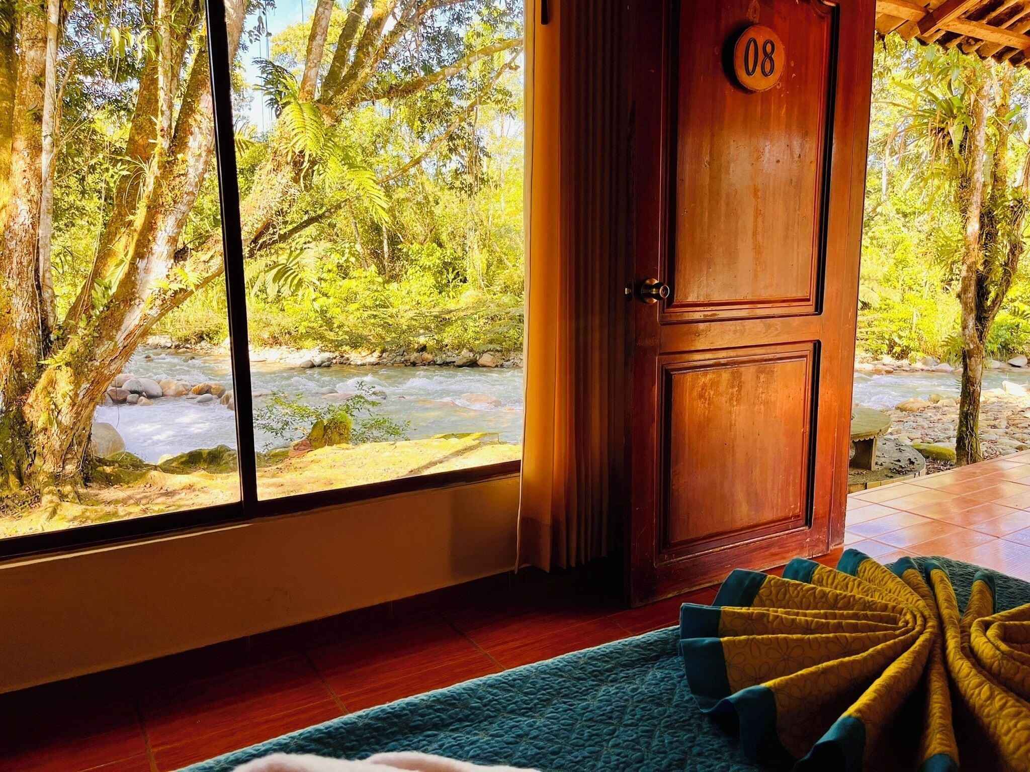 Una habitación con un gran ventanal con vistas a un río, una puerta de madera con un número y una alfombra de diseño azul y amarillo.