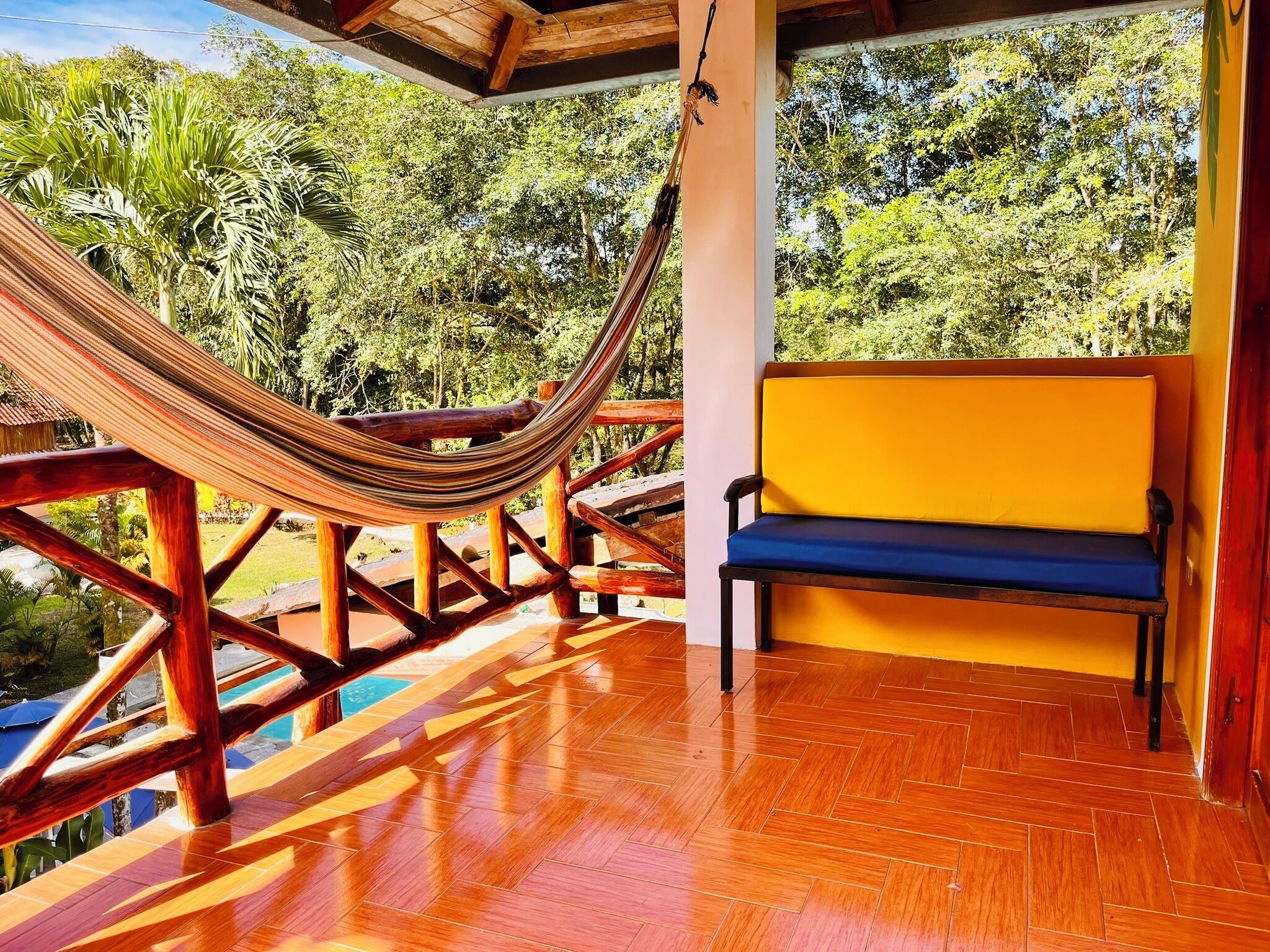 A hammock suspended between wooden beams, a bench with a blue cushion, and a wooden floor with a herringbone pattern.