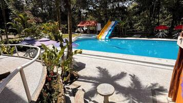 A swimming pool with a slide, surrounded by palm trees and a garden area with a stone bench.