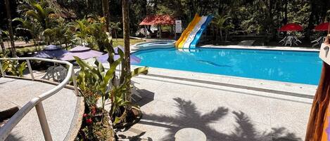A swimming pool with a slide, surrounded by palm trees and a garden area with a stone bench.