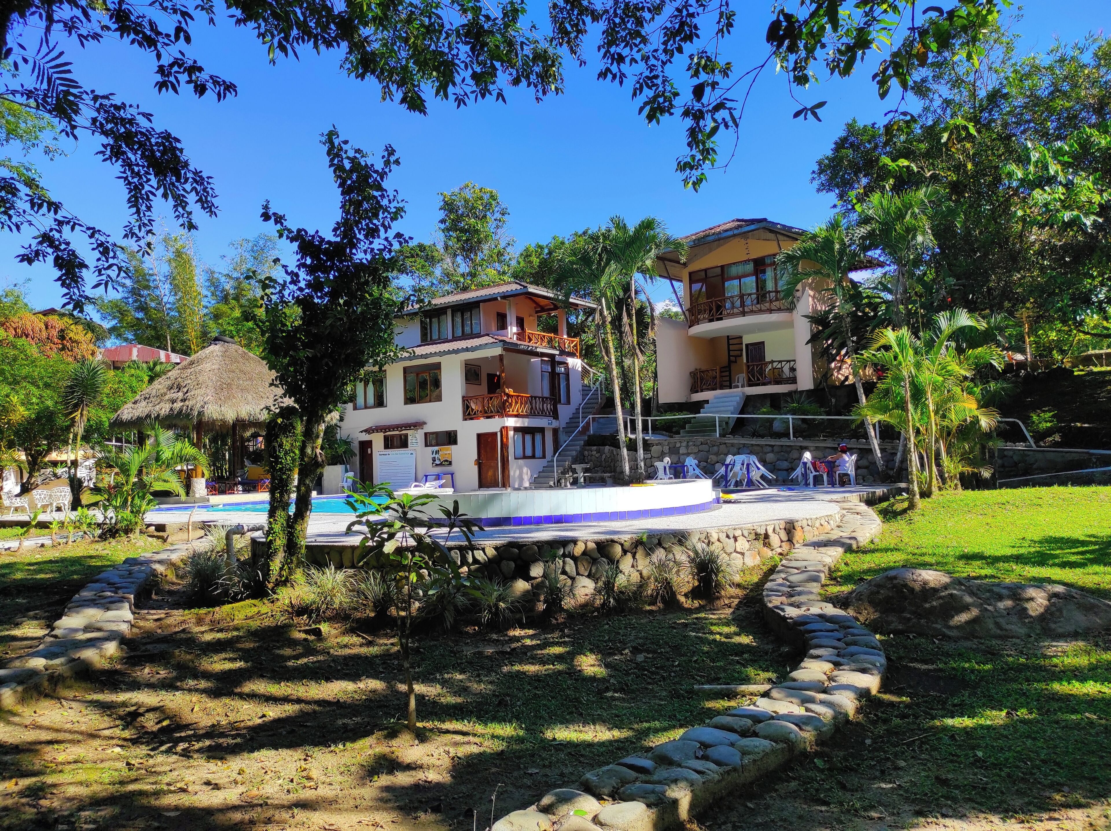 A two-story building with a pool, surrounded by greenery and a stone pathway.