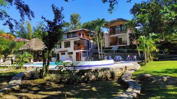 Un edificio de dos pisos con piscina, rodeado de vegetación y un sendero de piedra.