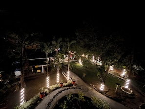 Vista nocturna de un jardín ajardinado con senderos, plantas en macetas e iluminación ambiental.