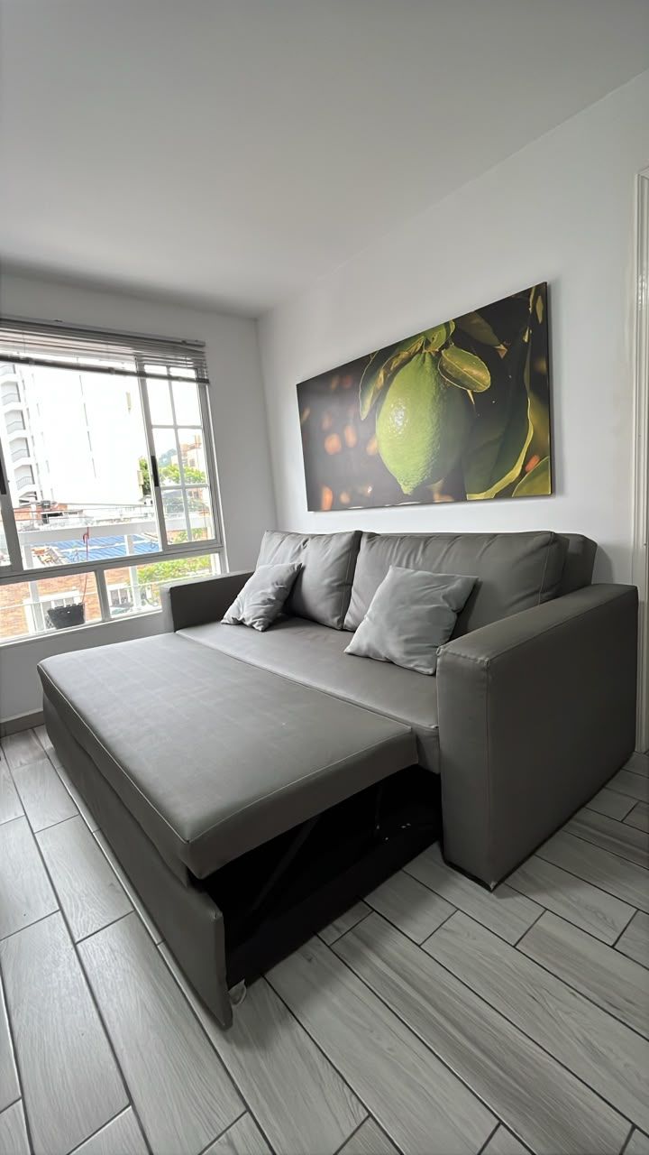 A modern bedroom with a grey sofa bed, a large window with blinds, and a city view.