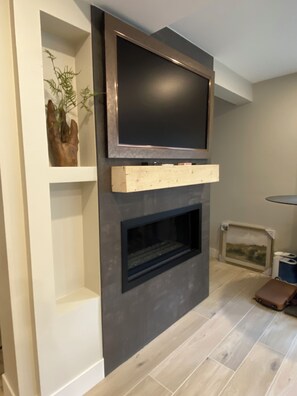A modern living room with a flat-screen TV mounted above a fireplace, a wooden shelf, and a framed picture on the wall. - Luxury 3BR/3BA Basecamp Steps to Quicksilver Lift Main Street Dining Heated Pool (Breckenridge)