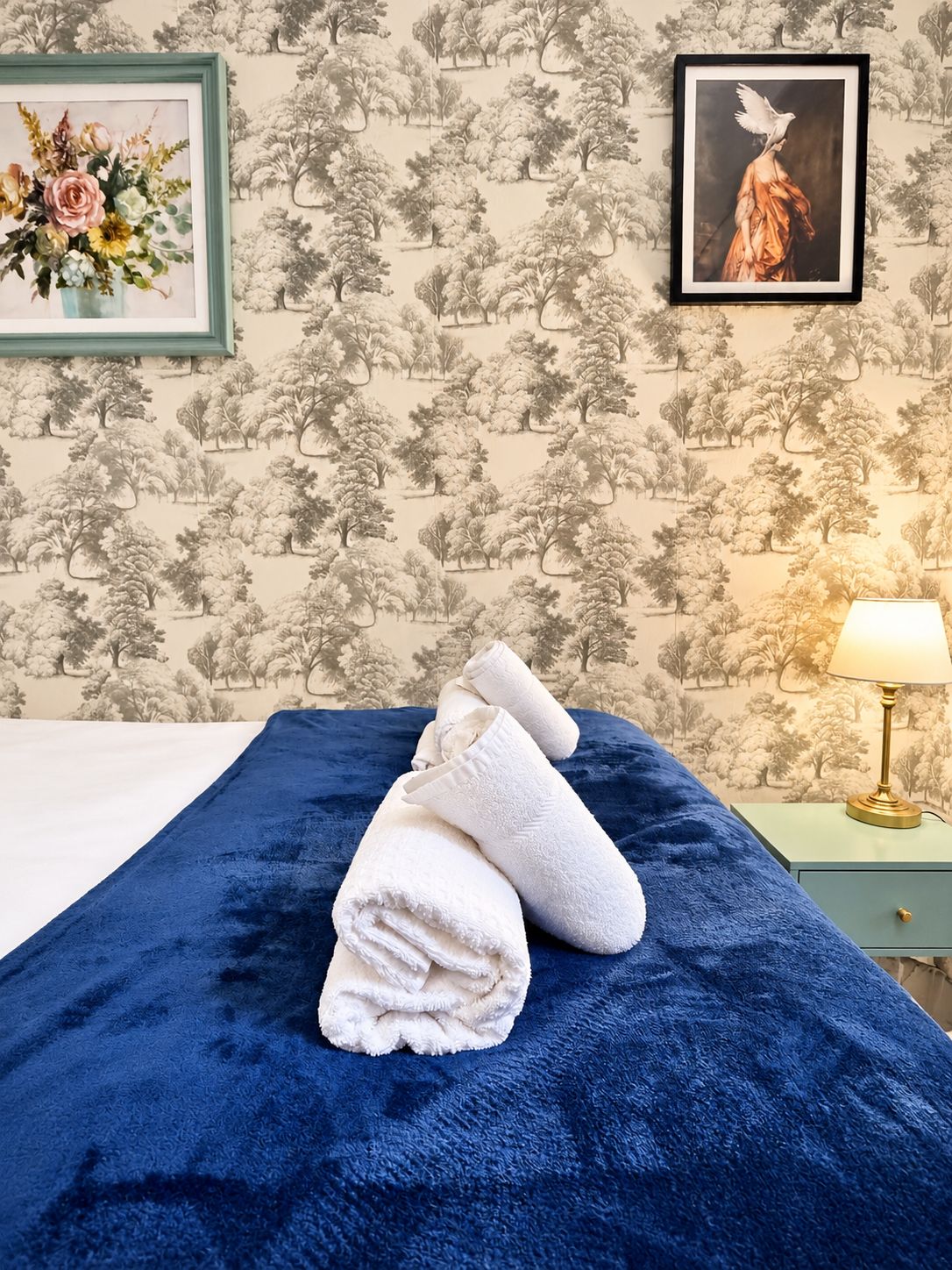 Ein ordentlich bezogenes Bett mit blauer Decke und aufgerollten weißen Handtüchern, ein gerahmtes Blumenbild an der Wand und eine Lampe auf einem Nachttisch.