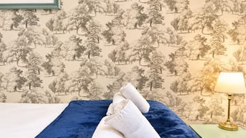 Ein ordentlich bezogenes Bett mit blauer Decke und aufgerollten weißen Handtüchern, ein gerahmtes Blumenbild an der Wand und eine Lampe auf einem Nachttisch.