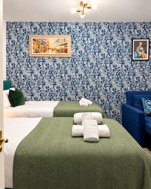 Ein Hotelzimmer mit zwei Betten, einer grün-weißen Bettdecke, einem blauen Sofa und einer Wand mit blumigem Tapetenmuster.