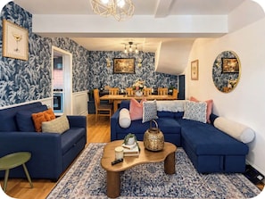 Ein Wohnzimmer mit einem blauen Sofa, einem Couchtisch und einem gemusterten Teppich. Im Hintergrund ist ein Essbereich mit einem Holztisch und Stühlen zu sehen.