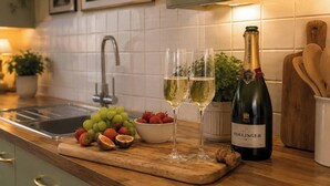 A kitchen counter with a bottle of Bollinger champagne, two glasses, a bowl of strawberries, and a pile of grapes.