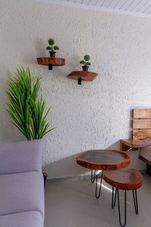 Ein Zimmer mit rauer, weißer Wand, zwei Holztische mit Metallbeinen, ein graues Sofa und zwei Wandbepflanzungen mit kleinen Bäumen.
