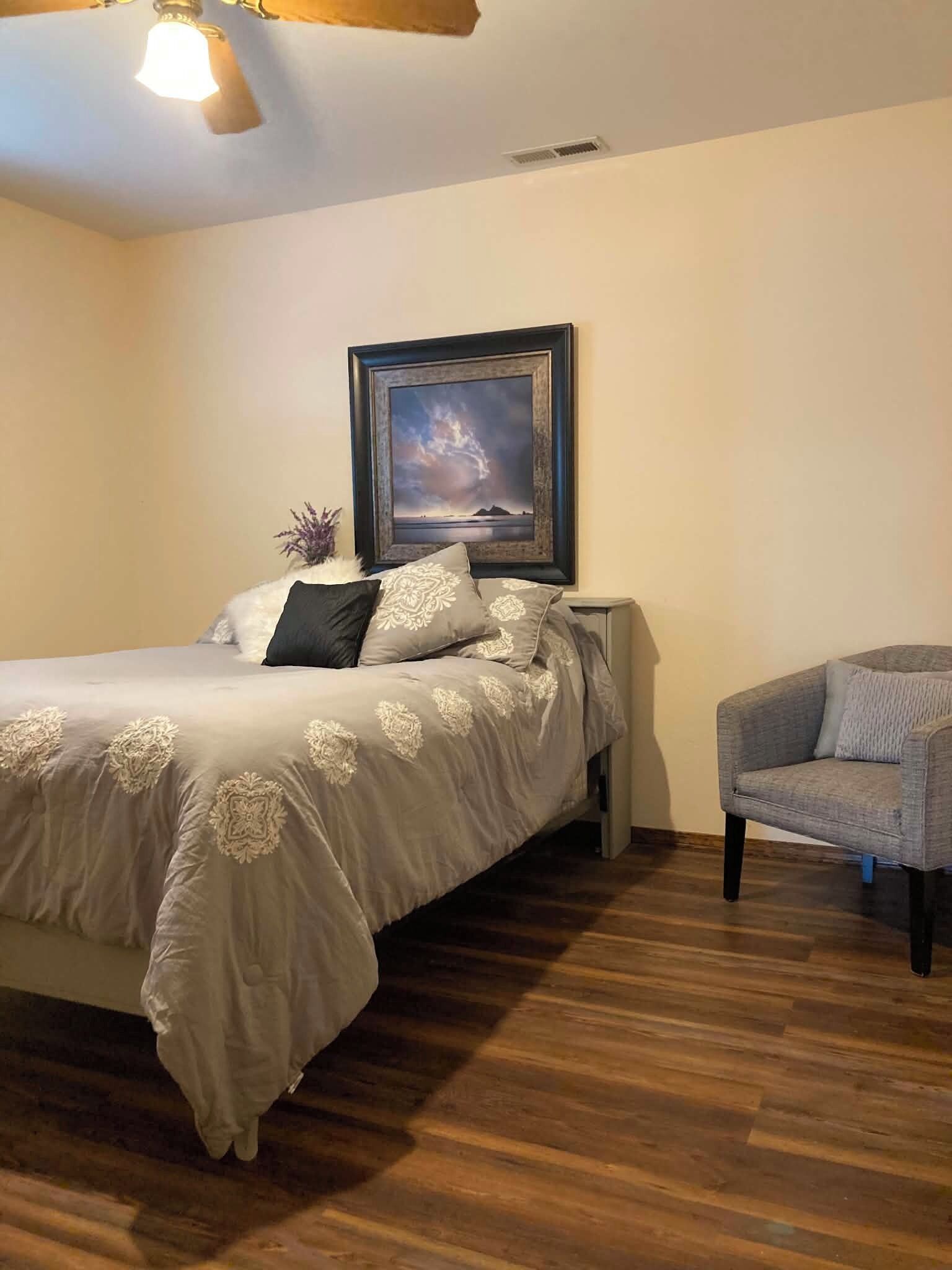 A bedroom with a bed, a chair, a framed picture on the wall, and a ceiling fan.
