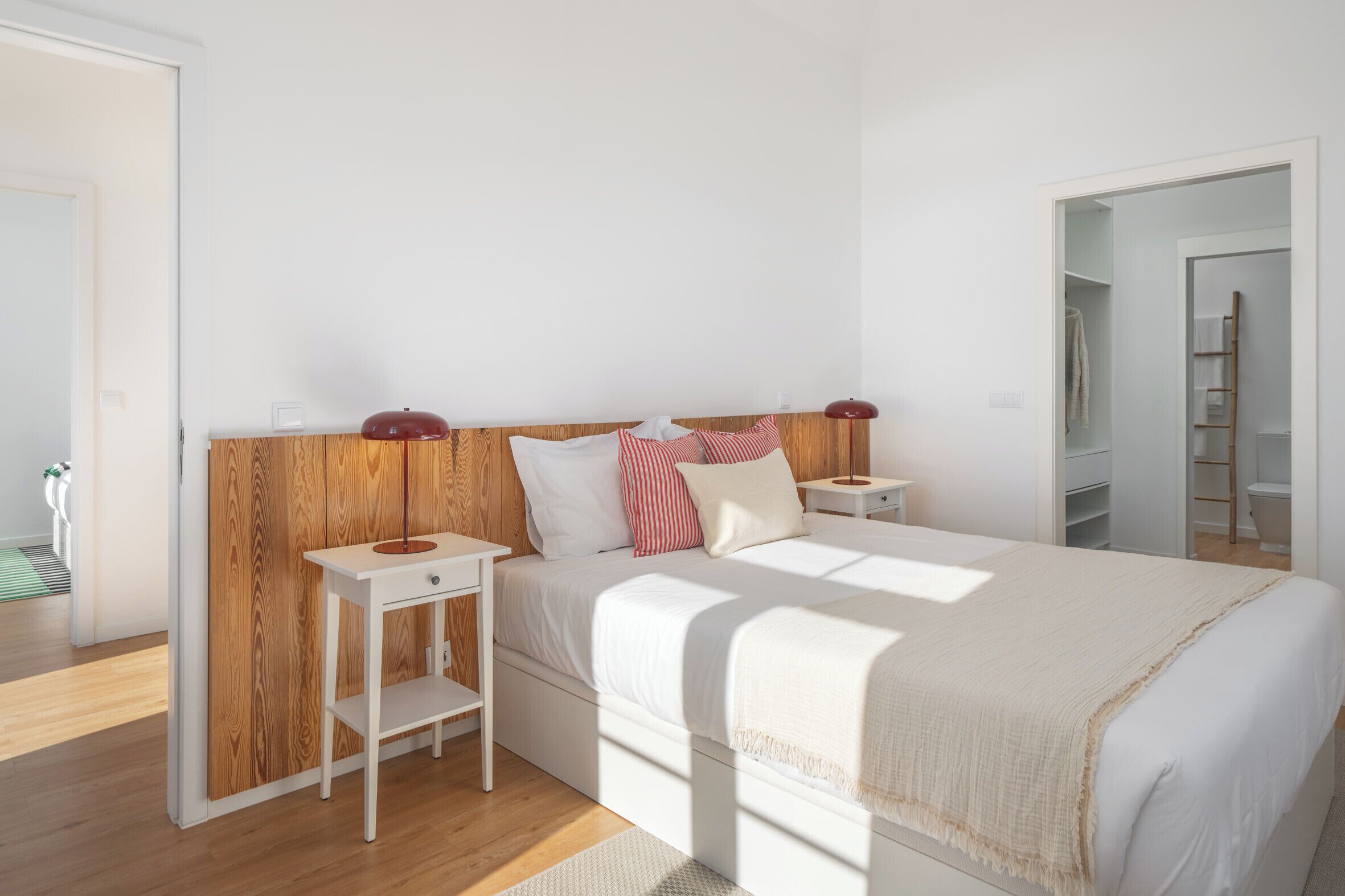 Quarto com cabeceira de madeira, cama com roupa de cama branca, duas mesas de cabeceira com abajures, e vista para um banheiro por uma porta aberta.