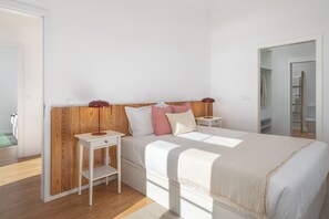 A bedroom with a wooden headboard, a bed with white bedding, two bedside tables with lamps, and a view of a bathroom through an open door. - Traditional House – Cozy 3-bedroom house (Funchal)