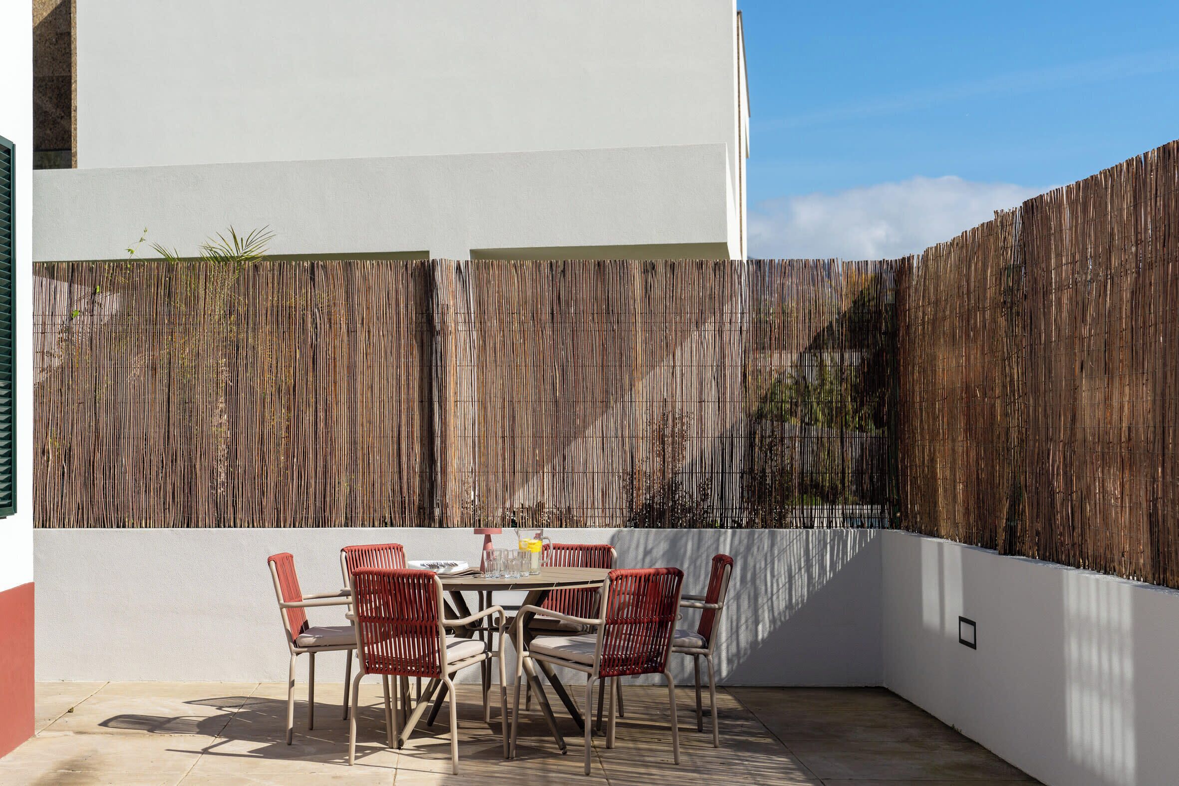 An outdoor patio with a table and chairs, a bamboo fence, and a white wall.