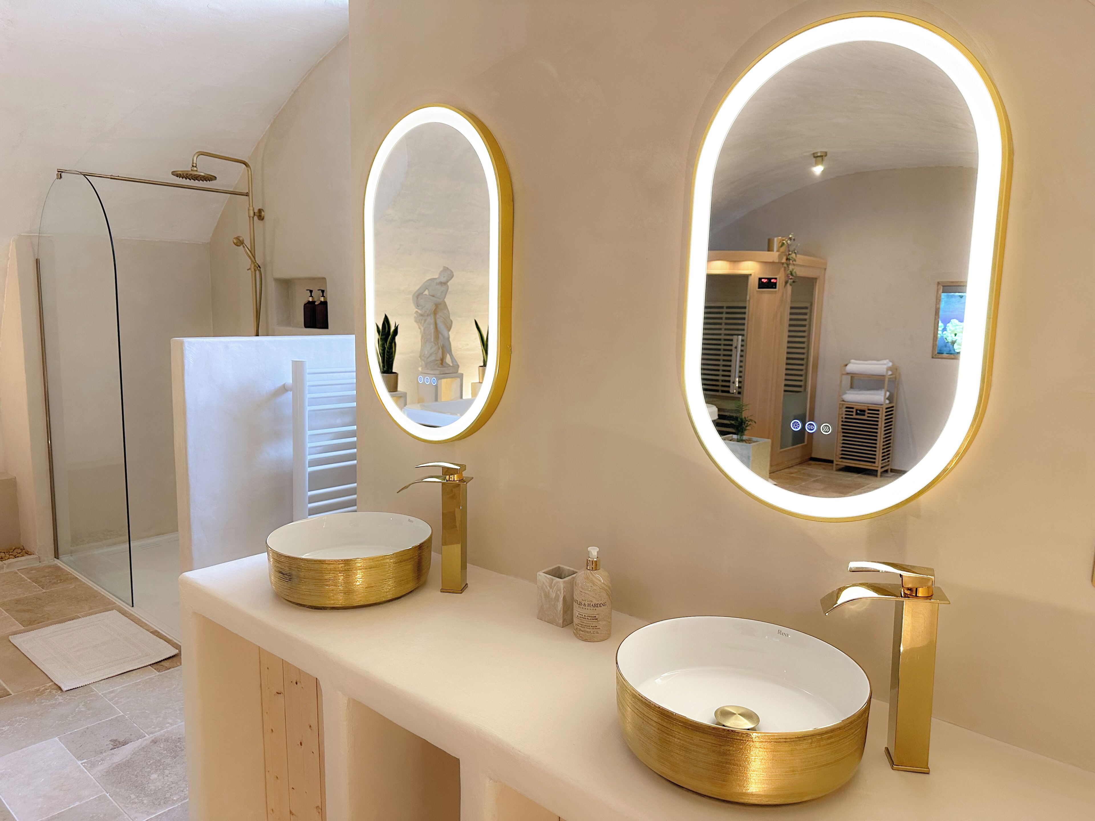 Un baño con dos lavamanos dorados, un espejo grande con iluminación y una zona de ducha.
