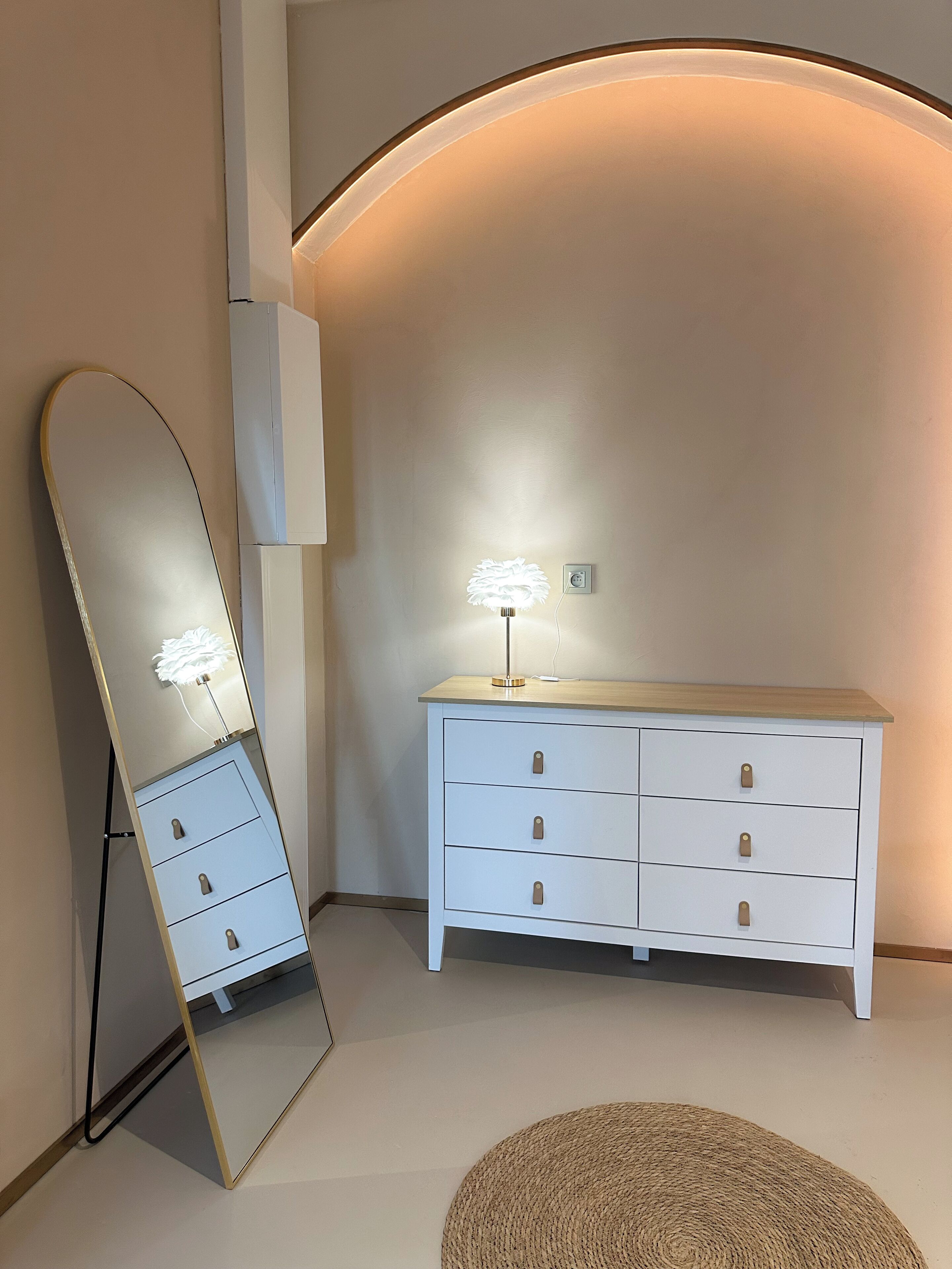 Ein Zimmer mit einem großen Spiegel, einer weißen Kommode mit Schubladen und einer Lampe.