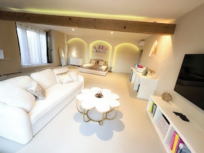A modern living room with a white sofa, a round coffee table, and a TV on a white stand. - Venus • Private Spa, Jacuzzi & Sauna (L'Arbresle)