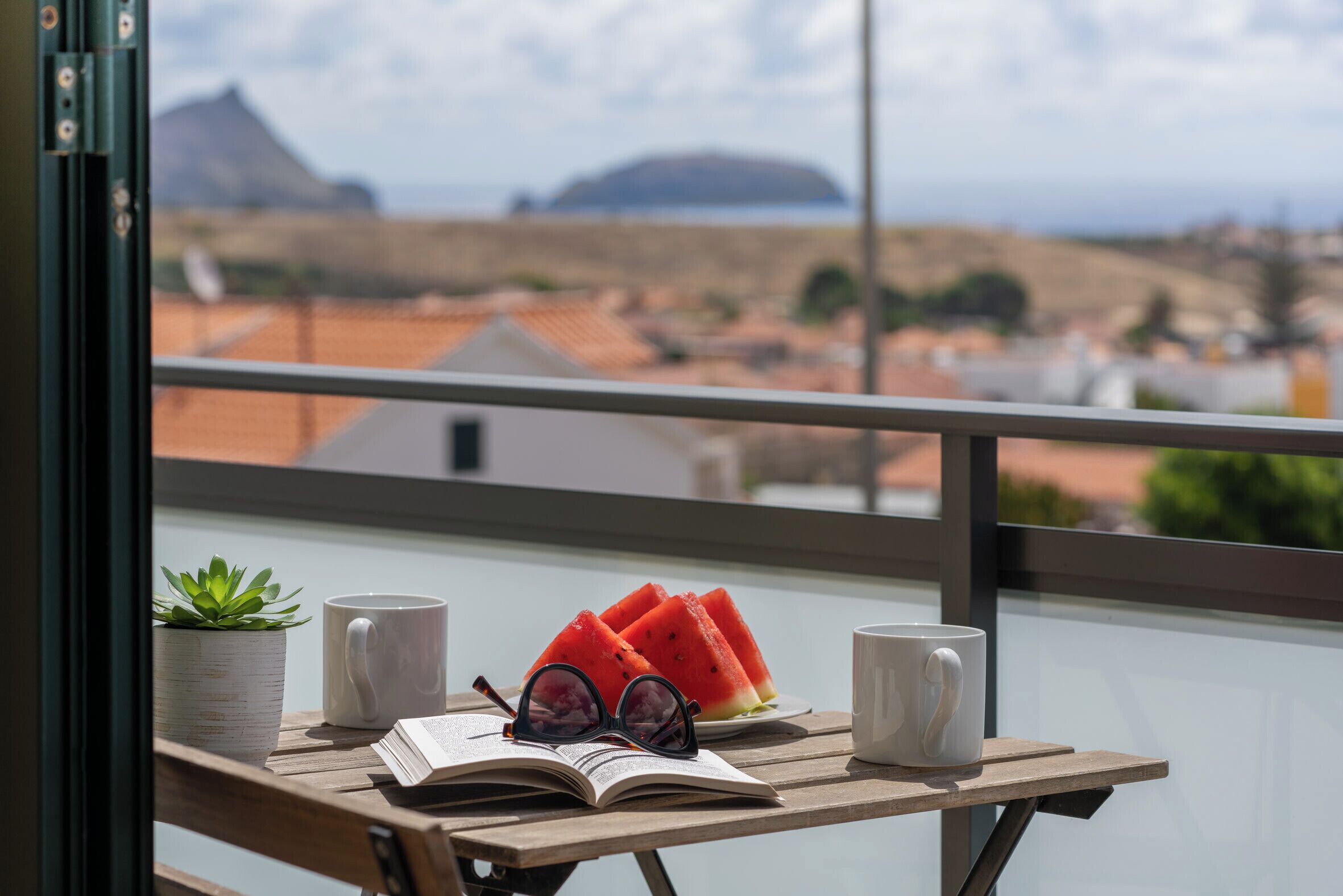Ein Balkon mit Holztisch, zwei Tassen, einem Buch, einer Brille und einem Blumentopf.