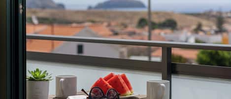 A balcony with a wooden table, two mugs, a book, a pair of glasses, and a potted plant.