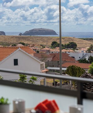 A coastal town with terracotta-roofed buildings, a clear view of the sea, and a prominent hill in the distance.
