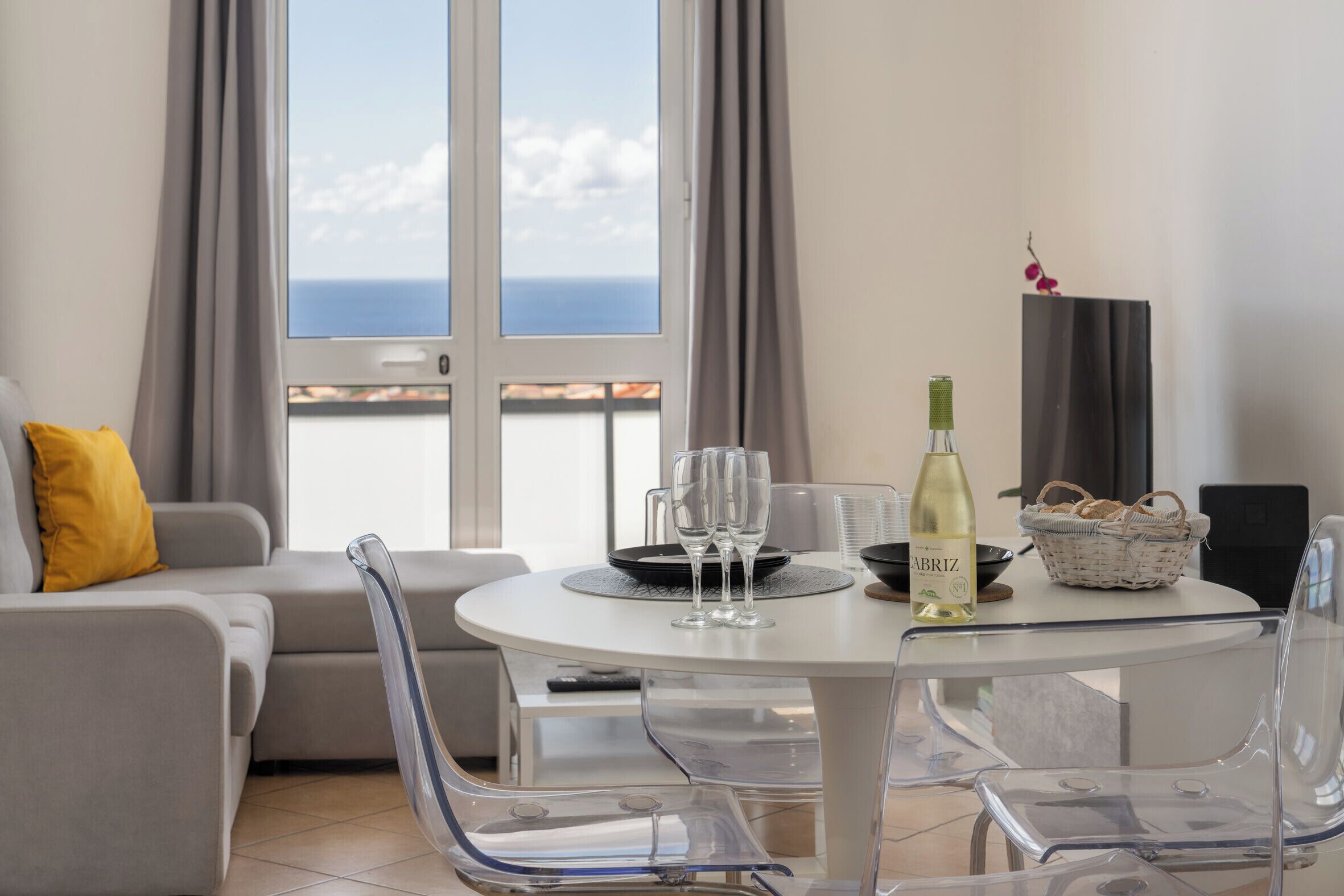Ein modernes Wohnzimmer mit einem runden Tisch, transparenten Stühlen, einem Sofa mit gelber Kissen, einer Weinflasche, Gläsern, einem Korb mit Brot und Meerblick durch große Fenster.