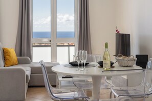 Un saló modern amb una taula rodona, cadires transparents, un sofà amb un coixí groc, una ampolla de vi, gots, un cistell amb pa i vista al mar a través de grans finestres.