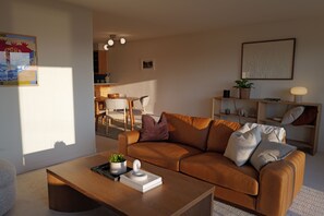 Una sala de estar moderna con un sofá de cuero marrón, una mesa de centro de madera con un libro y una planta, y un área de comedor al fondo.