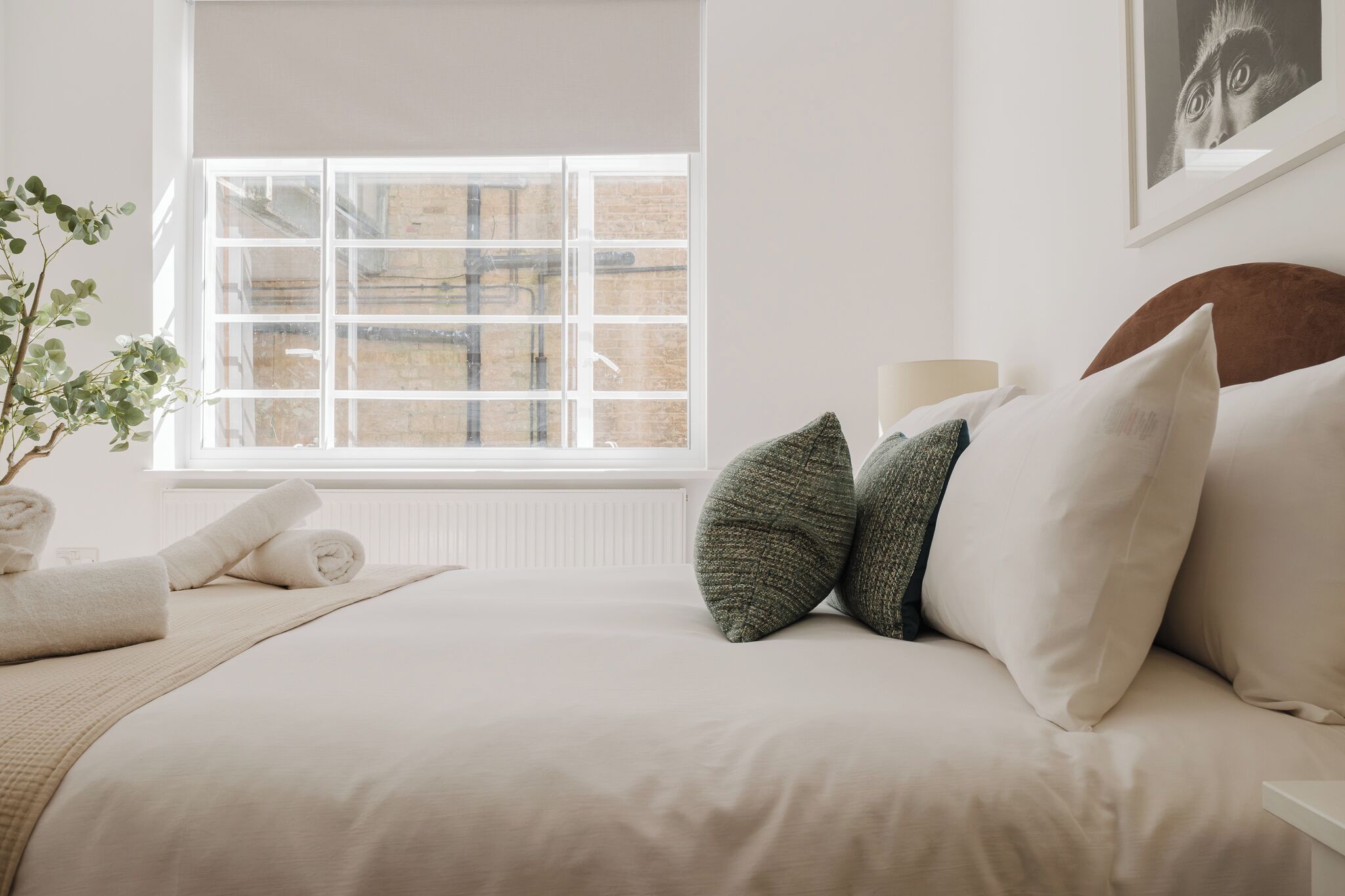 A neatly made bed with white bedding, a window with a view of a brick building, and a potted plant on the left.