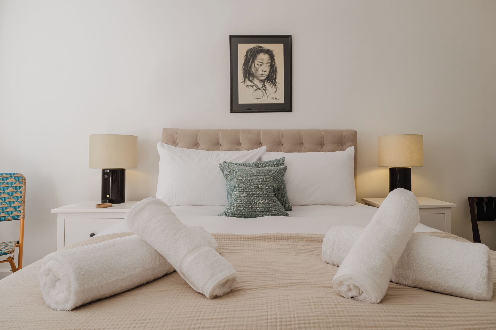 Una cama bien hecha con sábanas blancas, un cojín verde con textura y dos toallas blancas enrolladas. Hay dos lámparas de noche a ambos lados de la cama.
