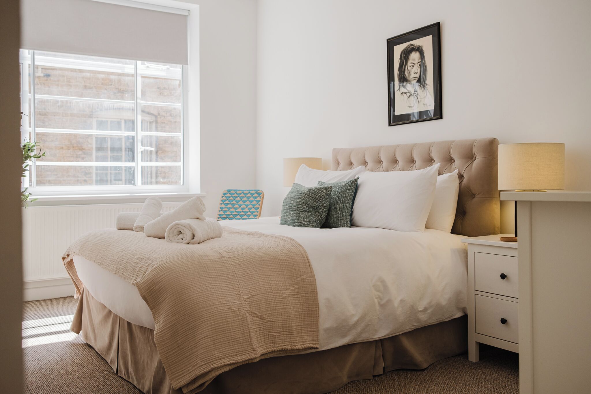 Un lit bien fait, avec une couette beige, des oreillers et une serviette enroulée. On aperçoit une table de chevet avec une lampe et un tableau encadré accroché au mur.