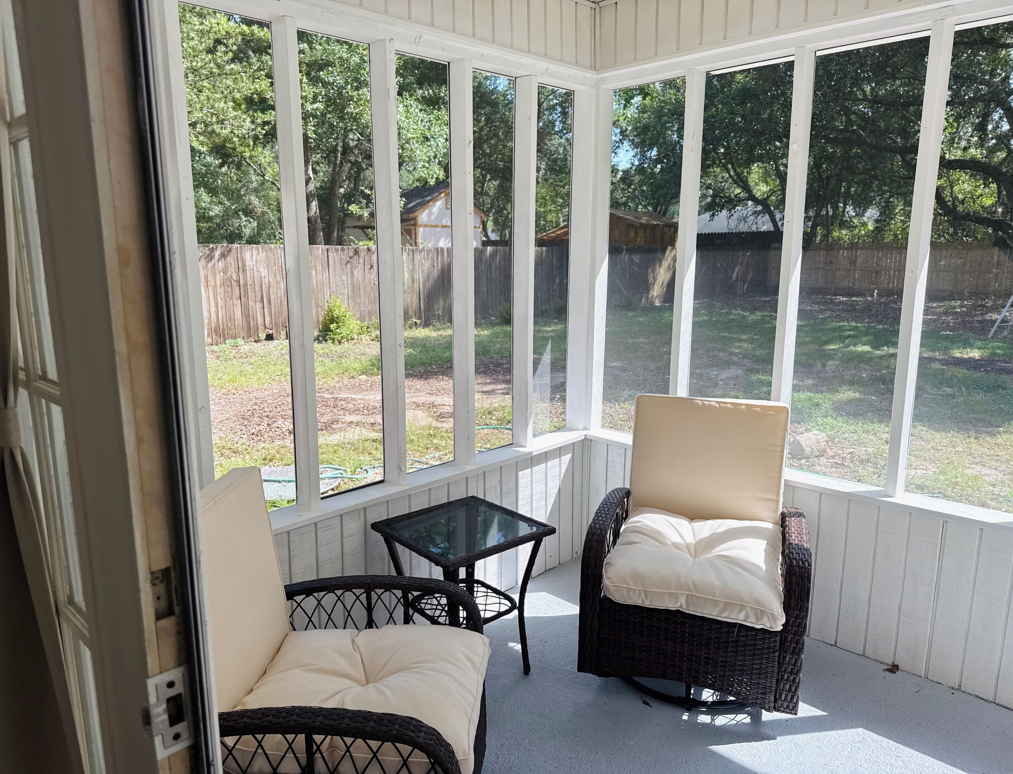 Ein überdachter Patio-Bereich mit zwei Korbstühlen und einem Glastisch, mit Blick auf einen Garten mit Holzzaun und Bäumen.