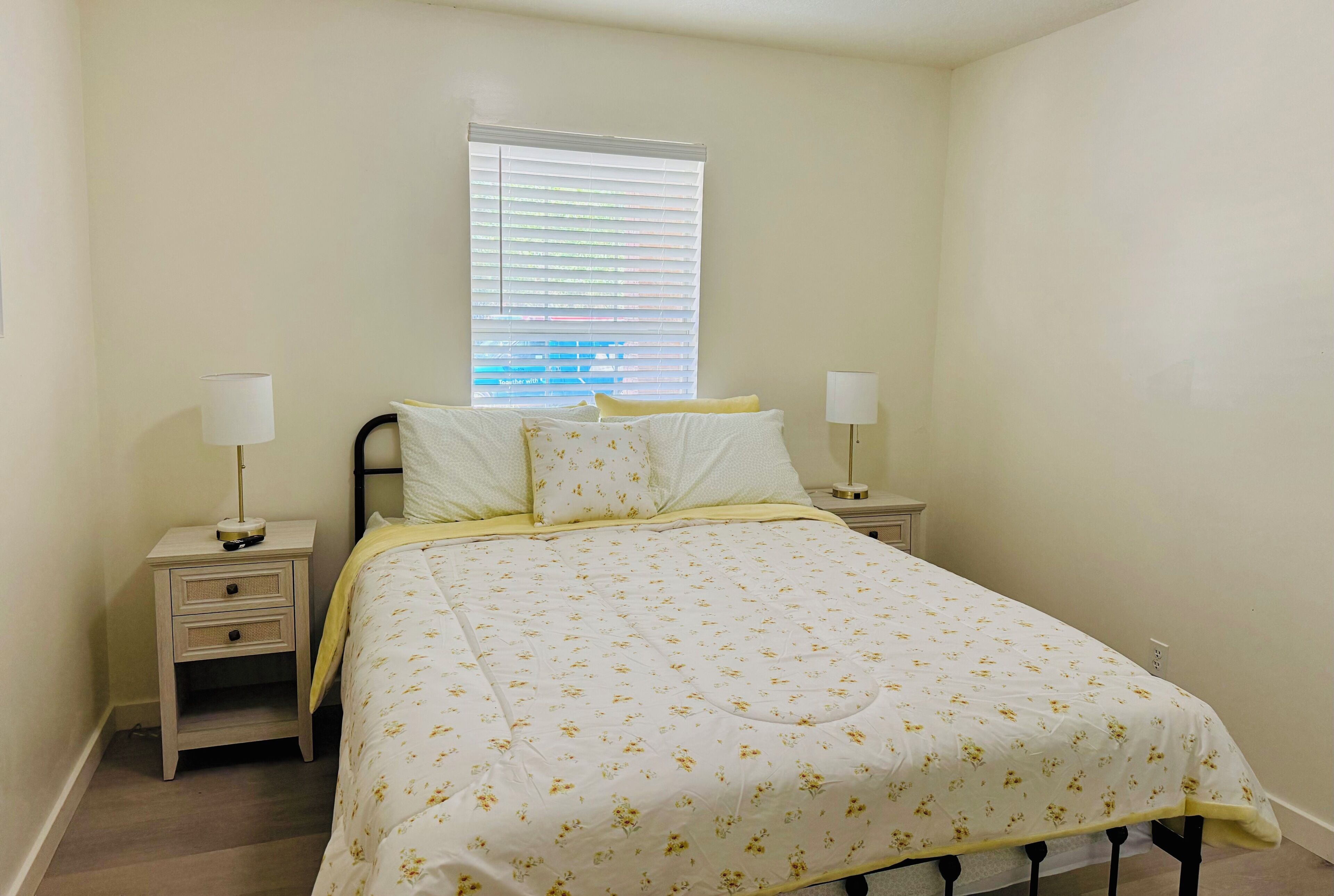 A bedroom with a bed, two bedside tables, a window with blinds, and a wall-mounted lamp.