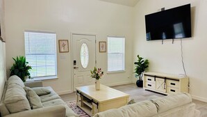 A living room with a beige sofa set, a coffee table with a vase, a flat-screen TV mounted on the wall, and a cabinet with drawers.