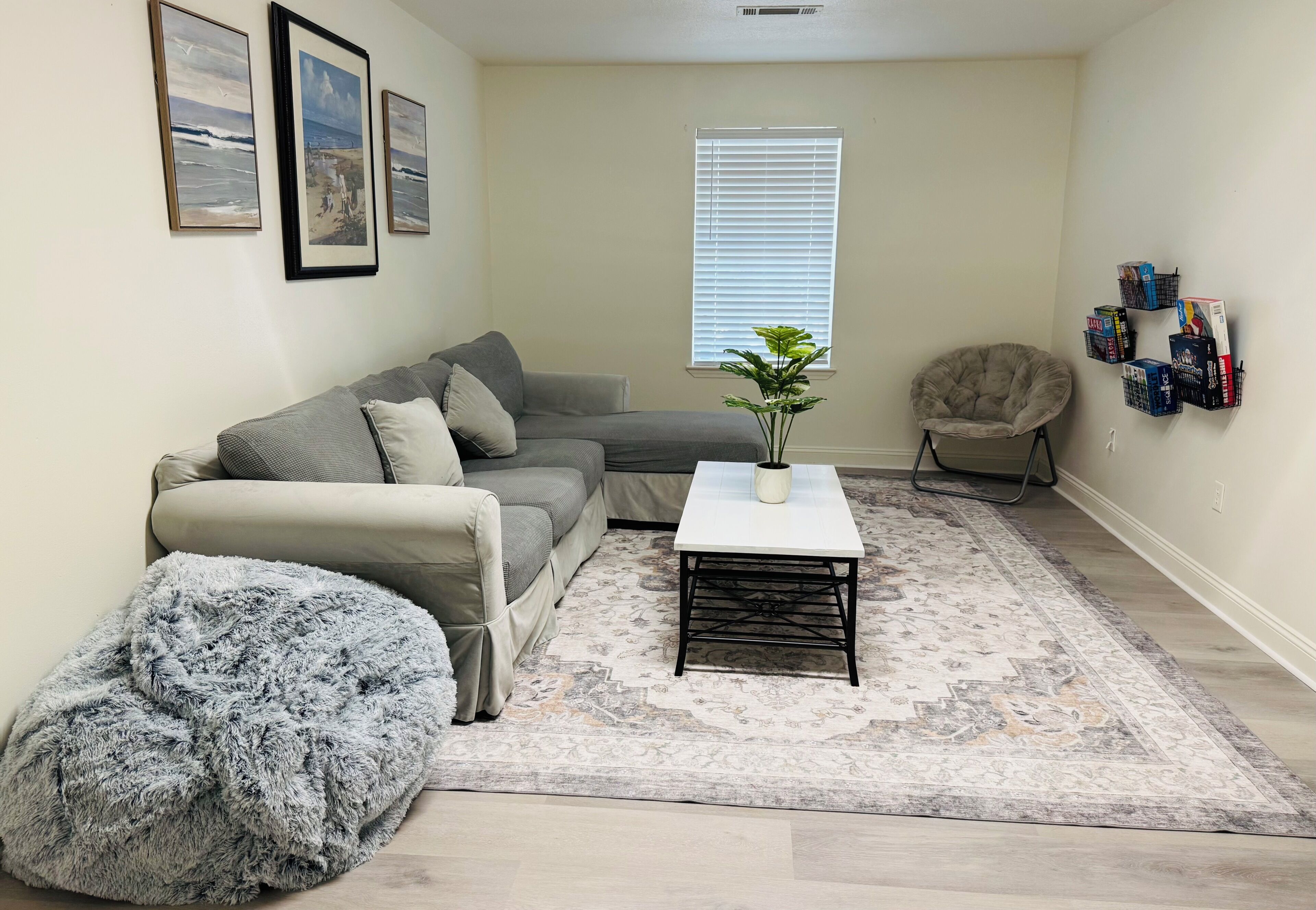Ein Wohnzimmer mit grauem Sofa, einem flauschigen Pouf, einem Couchtisch, einer Topfpflanze und einem Sessel.