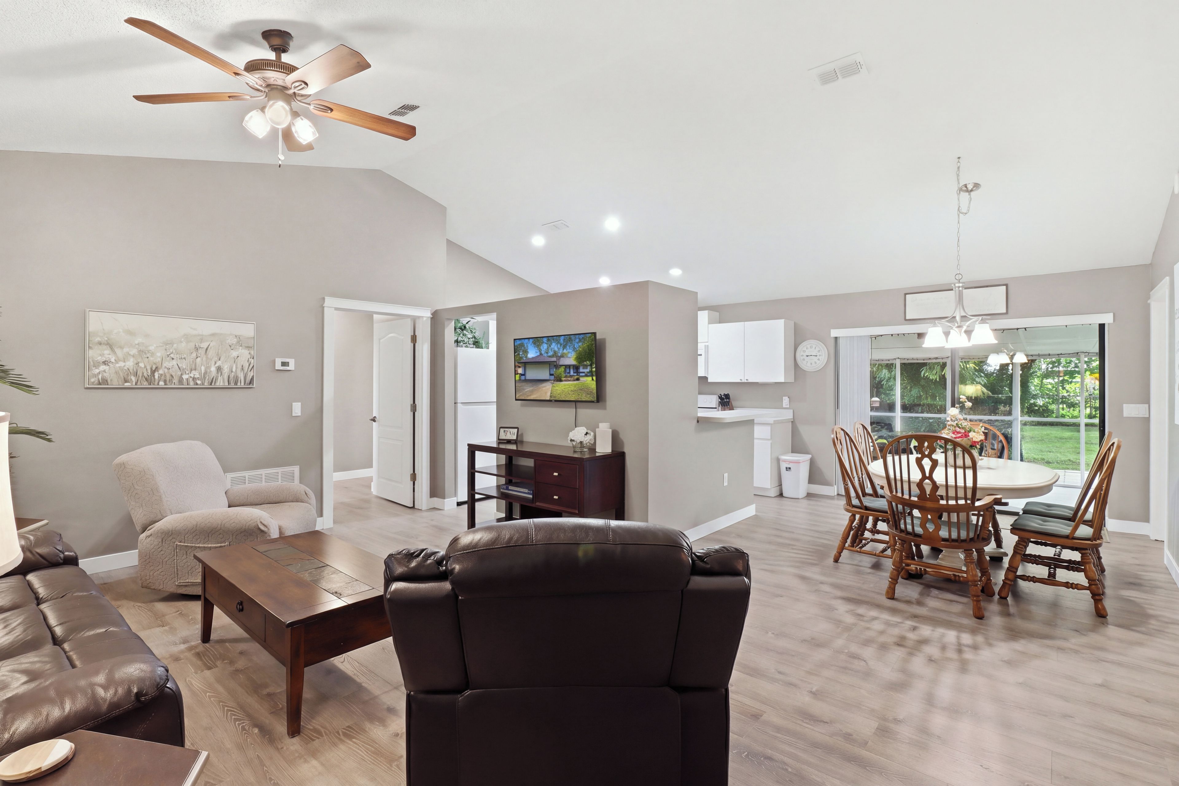 A spacious living room with a ceiling fan, a dining area with a table and chairs, a kitchen area, and a large window providing a view of the outdoors.