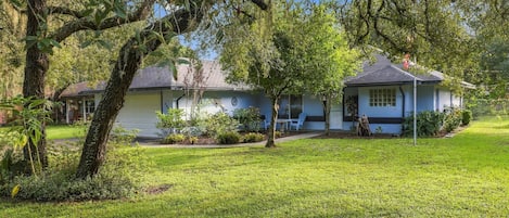 A residential house with a well-maintained lawn, a porch with outdoor seating, and a large tree in the foreground.
