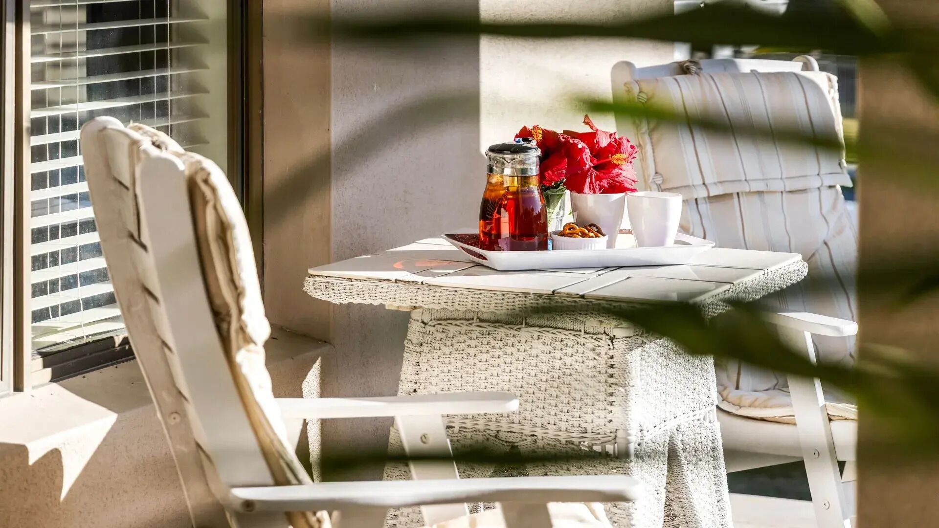 A wicker table with a white chair, a glass jar, a red flower, and a white cup.