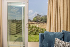 A room with a sliding glass door leading to a vineyard view, a blue sofa with a cushion, and beige curtains.