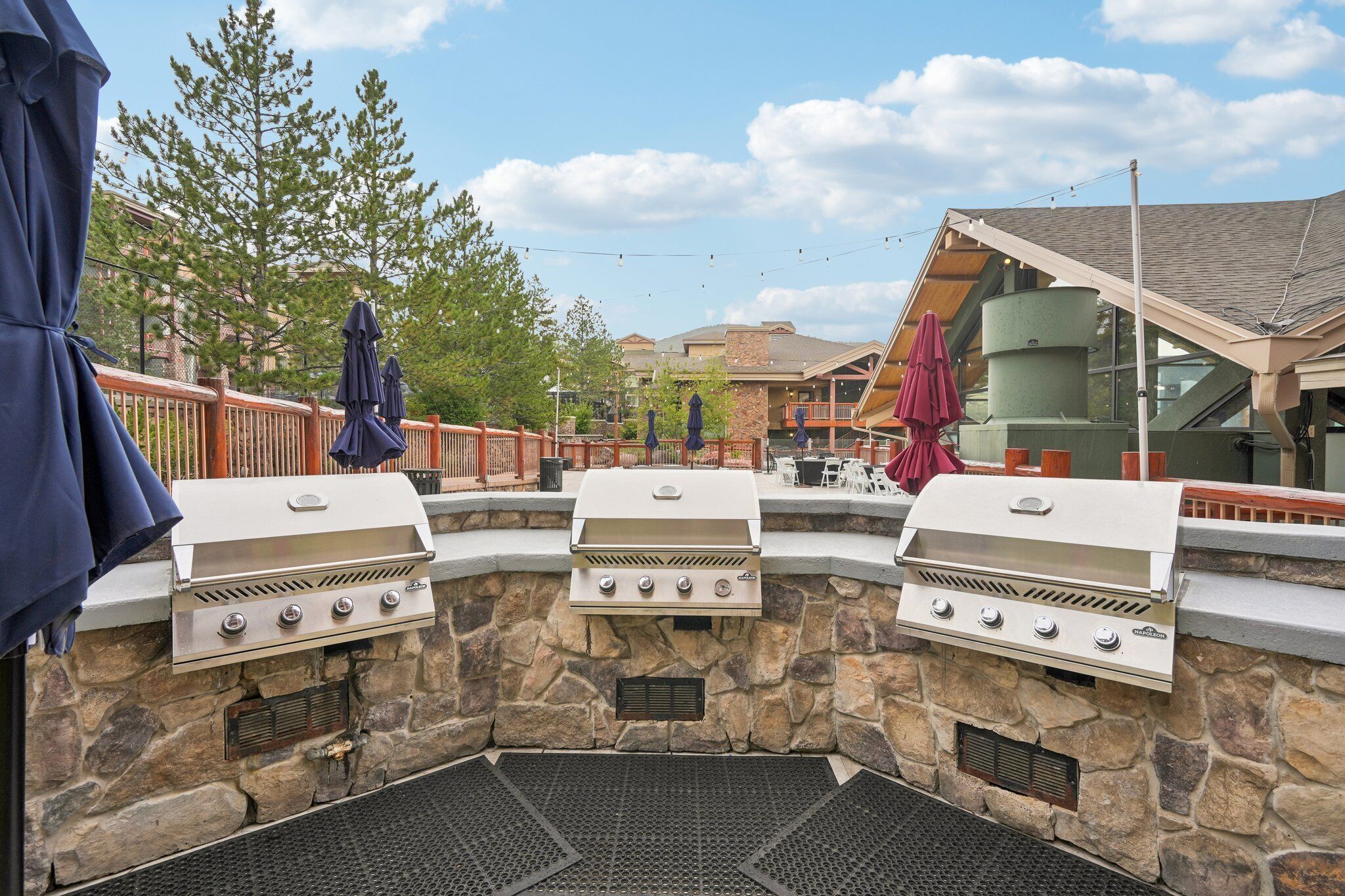 A stone barbecue area with three grills, outdoor seating, and a view of a building with a sloped roof.