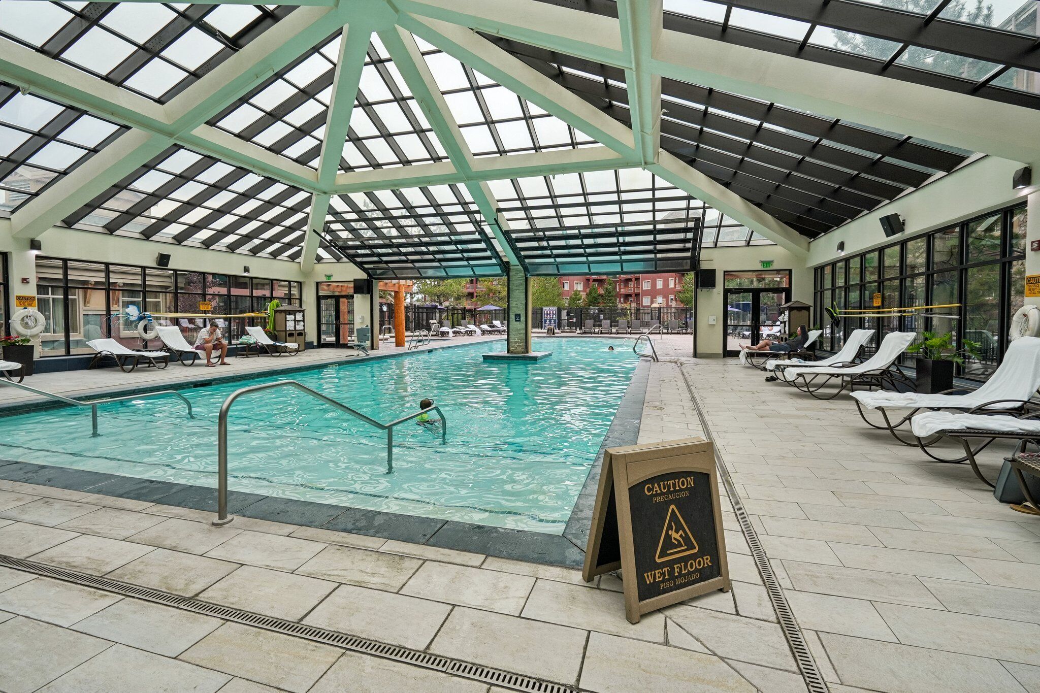 Ein Innenpool mit Glasdach, Liegestühlen und einem Warnschild wegen rutschiger Böden.