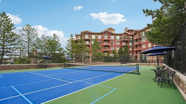 An outdoor tennis court with a net, surrounded by a stone wall, trees, and a building with balconies in the background. - Westgate Studio | Ski-In/Ski-Out | Pools, Spa & Ski Valet (Park City)