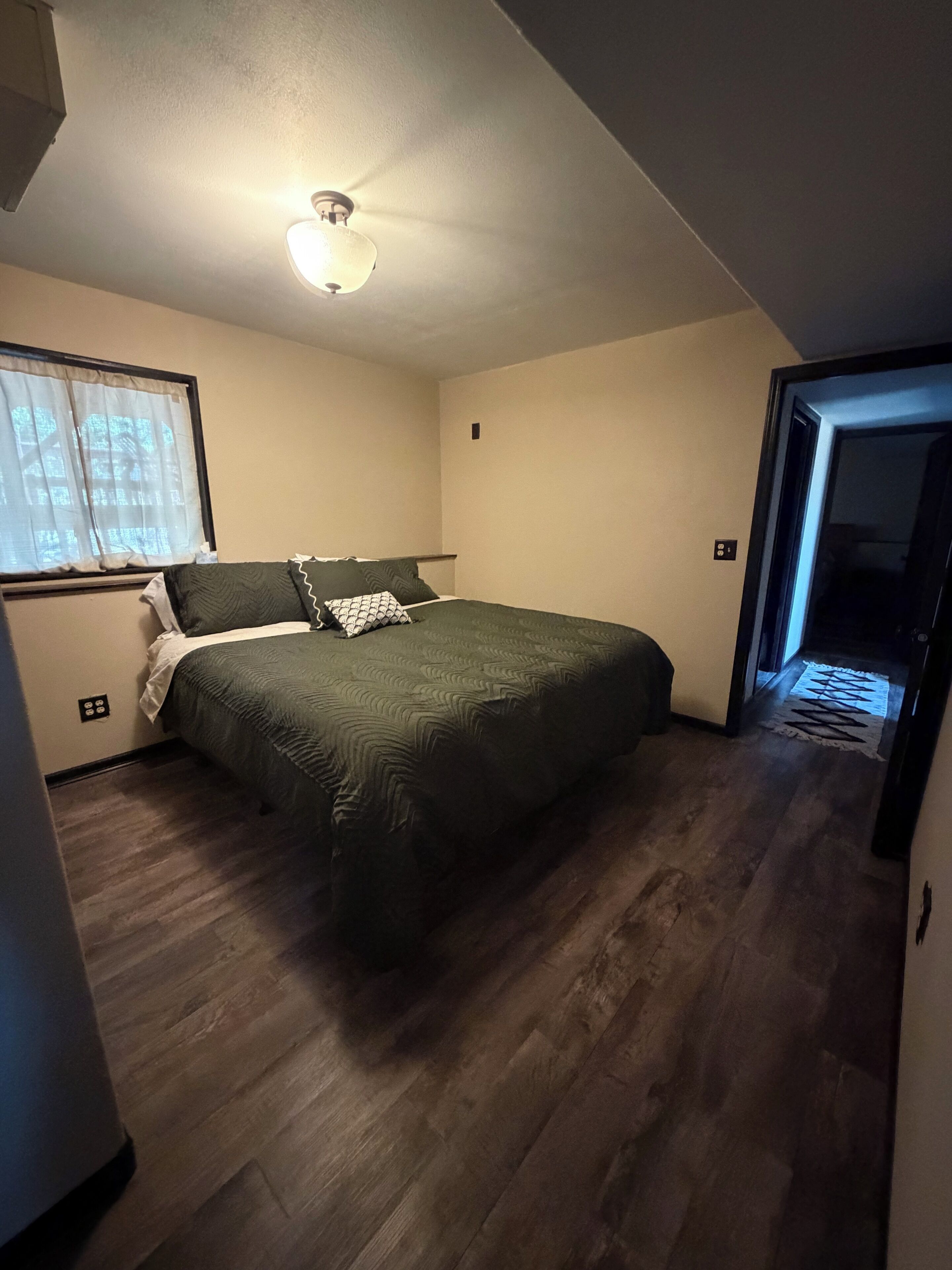 A bedroom with a bed, a ceiling light, a window with curtains, and a door leading to another room.