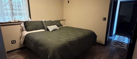 Ein Schlafzimmer mit Bett, Deckenleuchte, Fenster mit Vorhängen und einer Tür zu einem weiteren Raum.