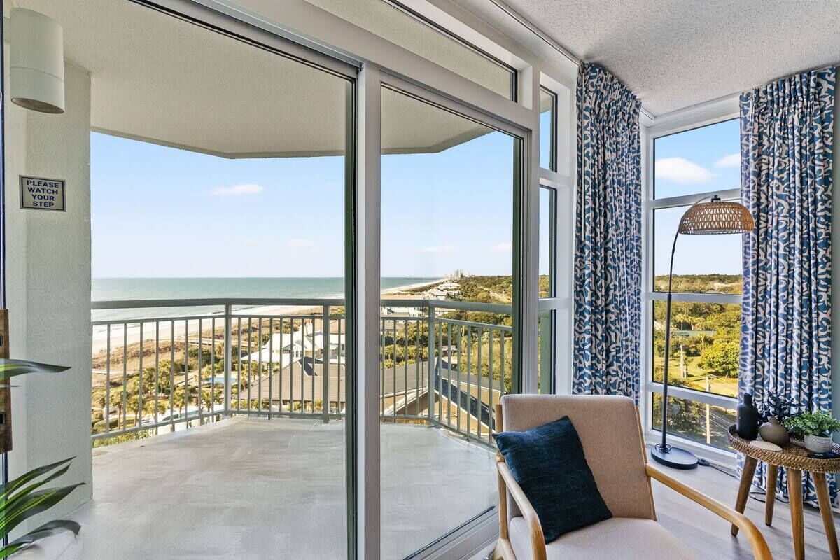 A balcony with a view of the beach and ocean, a chair with a cushion, and a floor lamp.