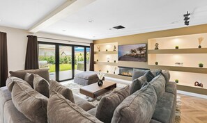 Ein modernes Wohnzimmer mit Kamin, einer großen Couch und einem Couchtisch. Ein großes Schiebeglasfenster führt auf einen Balkon.