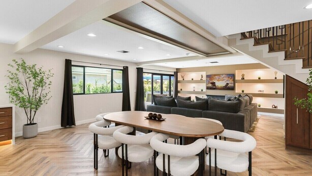 Uma sala de estar moderna com um sofá, uma mesa de jantar com cadeiras, uma escadaria e vista para o exterior.