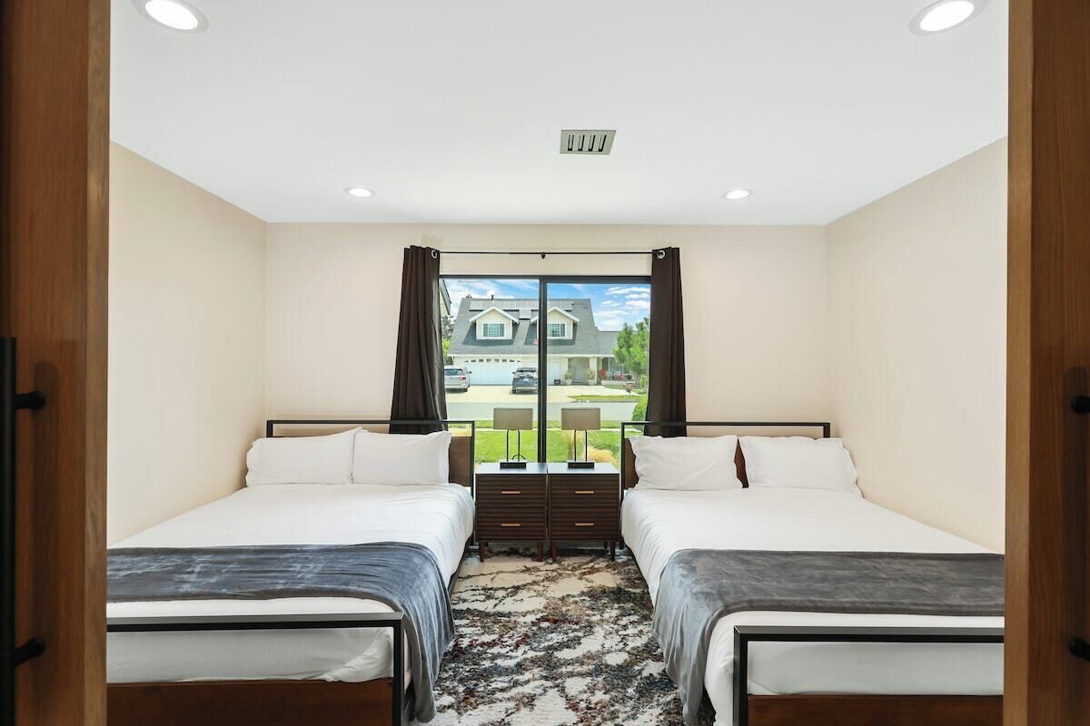 Une chambre avec deux lits, une grande fenêtre avec des rideaux, un luminaire au plafond et une vue sur une maison à l’extérieur.