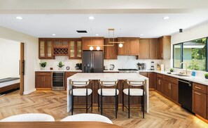 A modern kitchen with wooden cabinets, a central island with bar stools, and a large refrigerator. - Modern Luxury Designer Home (Midway City)